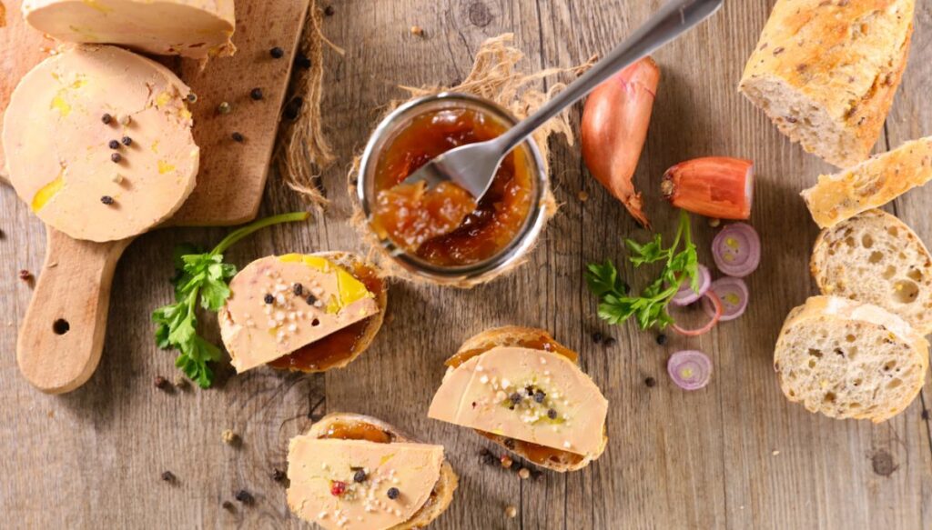 This ia an aerial view of a table with foie gras sitting on a cutting board on the left. In the middle you have a chutney in a bowl with a spoon and then foie gras on slices of baguette. On the right you have baguetta out and sitting on the table. 