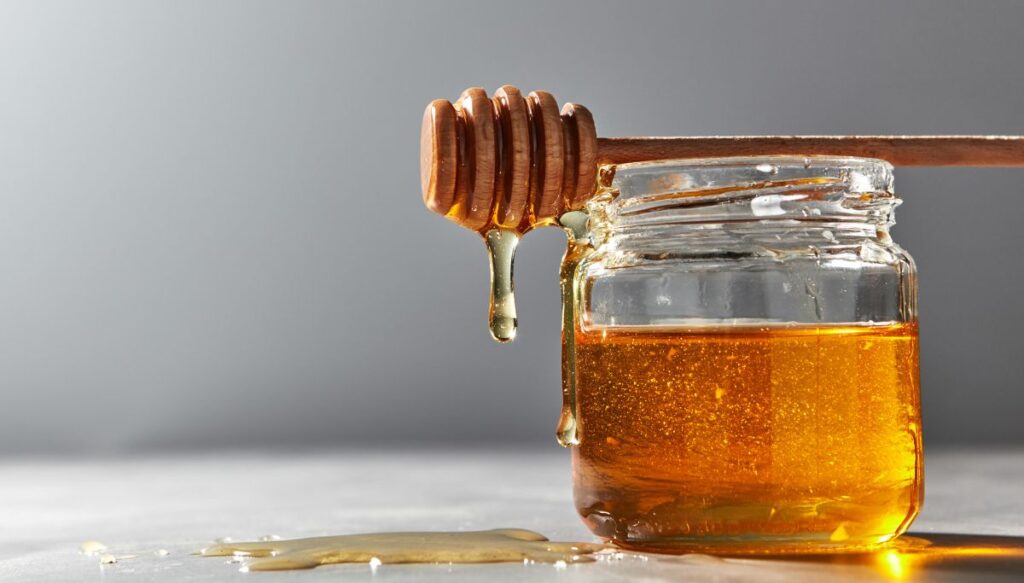 A view of a glass jar filled with honey. It sits on a table with honey dripping from a honey spoon that sits on top of the pot and has honey dripping from it with a gray wall in the background. Honey is a delicious tradtional food in Paris. 