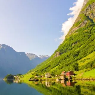 A view of Nærøyfjord with it's steep green cliffs and beautiful water. You can see a green mountain jutting out into the water with brown houses perched on the edge of the water.