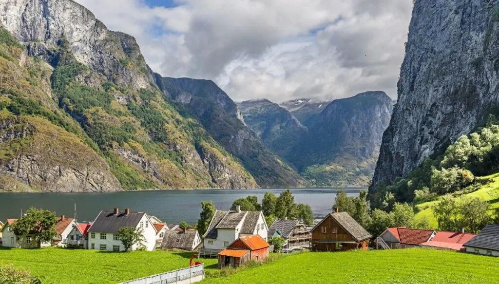 A view of the red. wood homes in the fjords of Norway. That sit on the green grass near the water and are surrounded by the mountains and fjords.