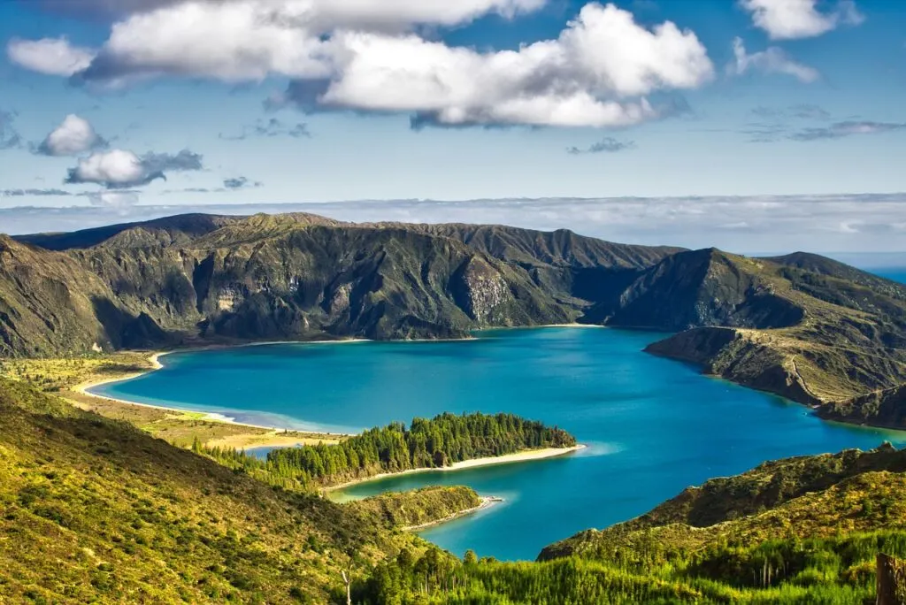View of a lake with bright blue water and rugged, forested mountains surrounding the lake on a sunny day in the Azores. 