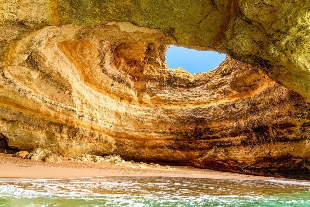 View of a white sand beach with blue water that is covered by a white stone cave with a giant hole in the roof. 