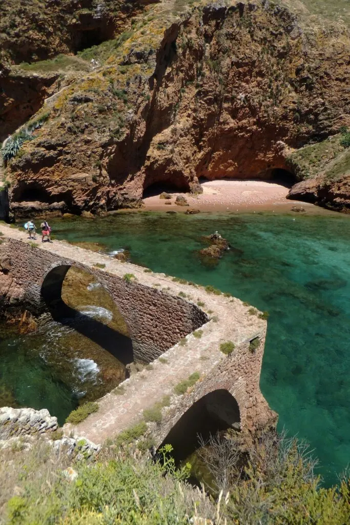 View of the steep cliffs that surround the beaches and blue waters of the Berlengas Islands. You can see people walking across an old stone bridge on a sunny day. 