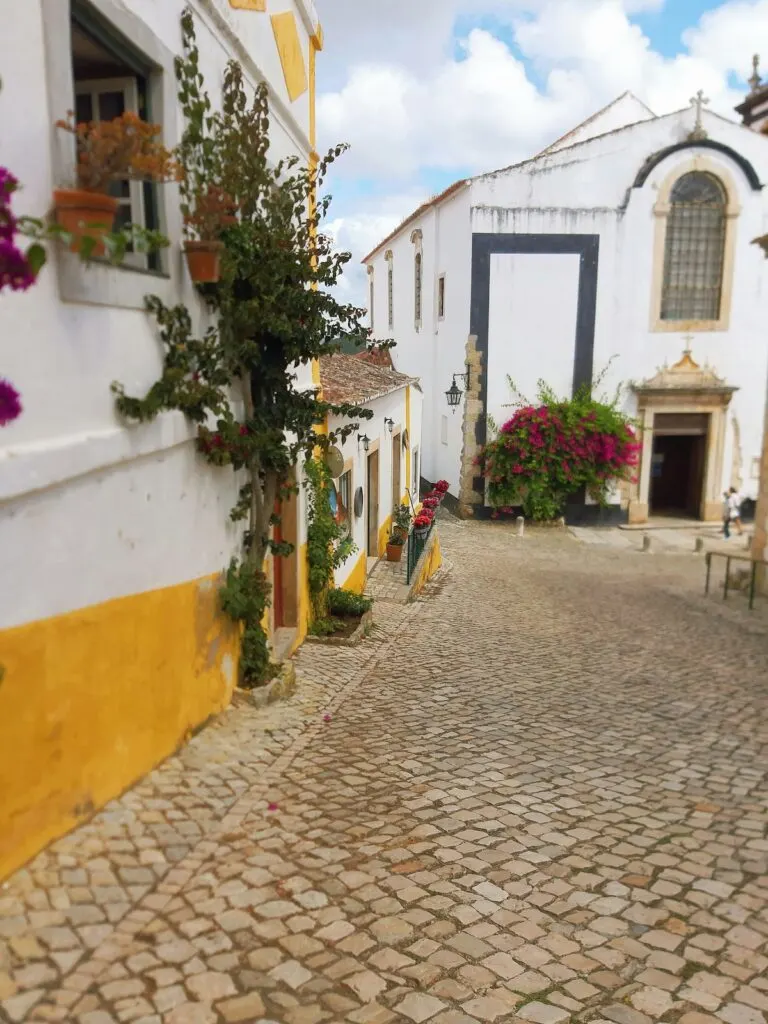 View of the white buildings with yellow trim and cobblestone streets that line the town of Obidos. You can see the church in the background as well. 