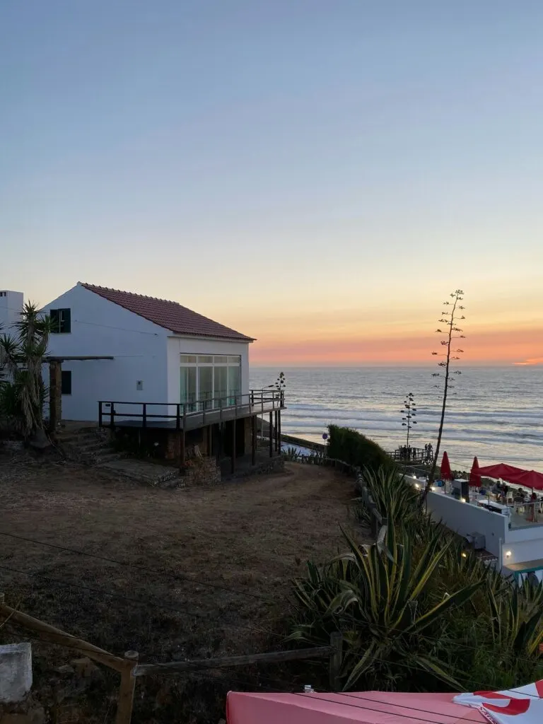 View of a white house with a porch looking out at the ocean and the sunset in Praia de Odeceixe. 