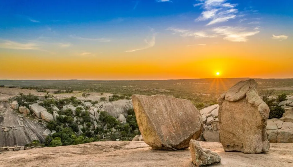 View of large rocks sitting on the ground in the foreground from the top of Enchanted Rock. You can see green plains and the sun setting in the sky. 