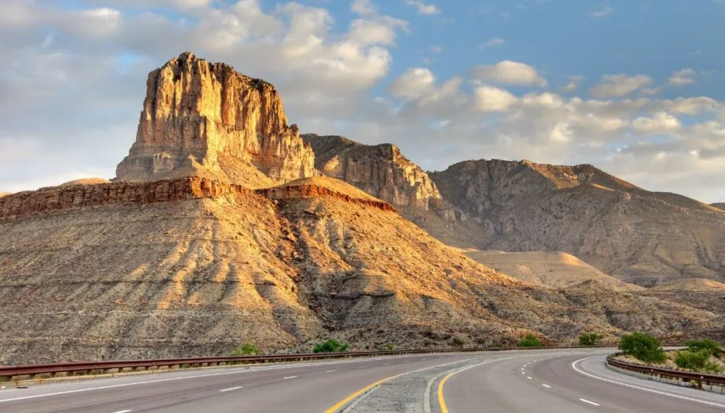 View of the striped mountains and uniqu rock formations that make up Guadalupe Mountains National Park. You can see a road through the park and clouds in the sky at sunset. 