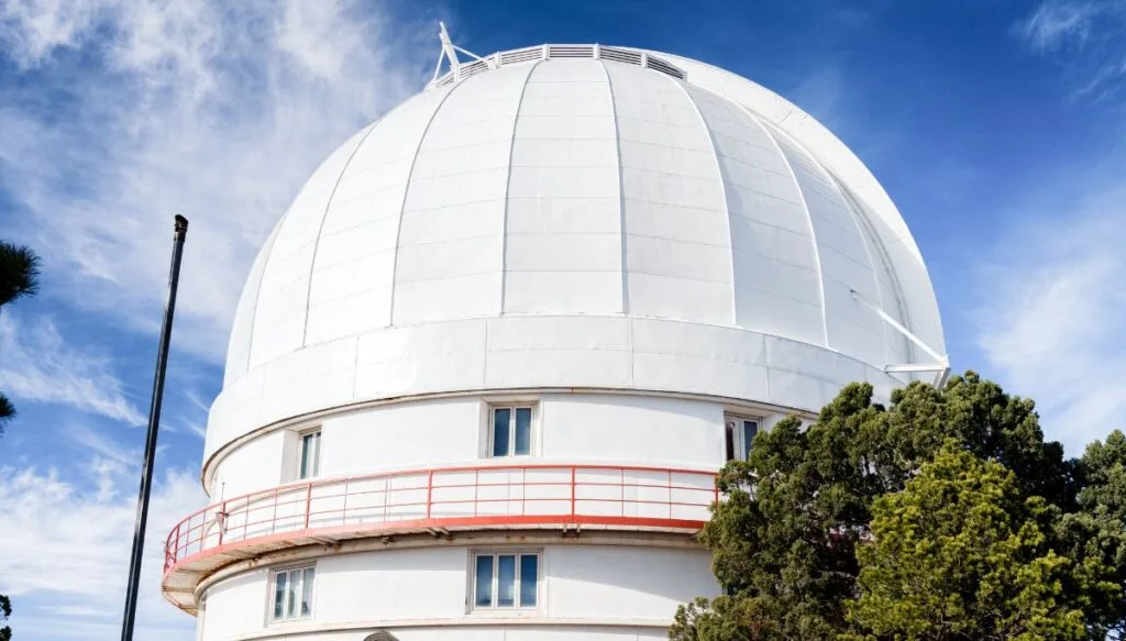 View of a white dome with windows and a pink railing and a cat walk around the edge with trees on the ground below. 