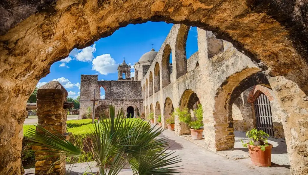 View through a stone arch of stone arches and towers with potted plants in the middle at Mission San Jose in San Antonio. 