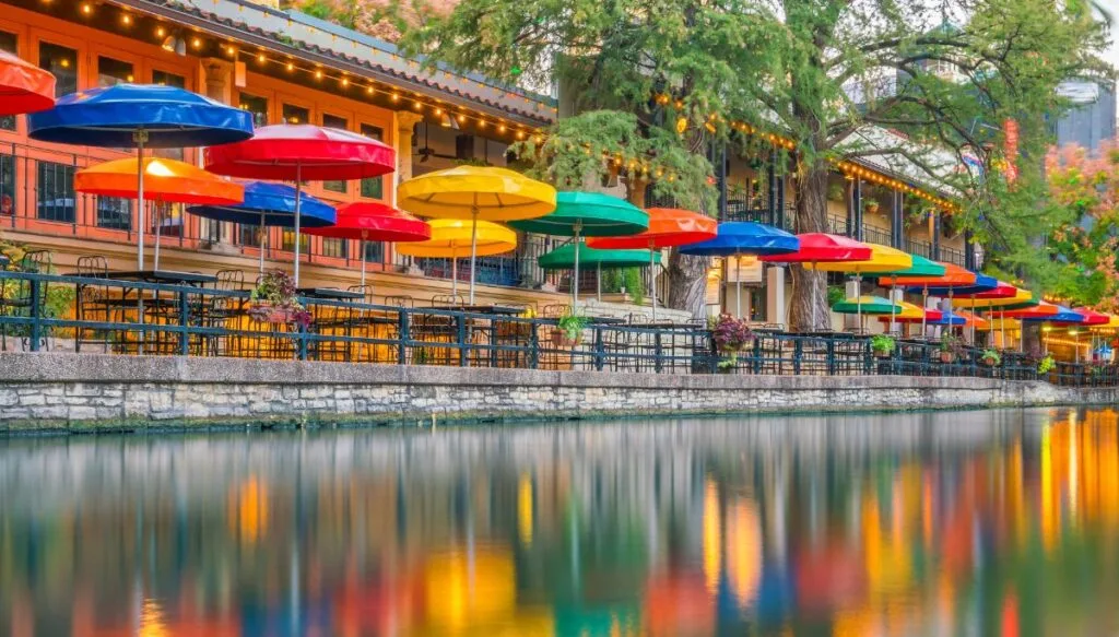 View from the river of all the blue red, yellow, green, and orange umbrellas that ;ine the pathway along the San Antonio River Walk. 