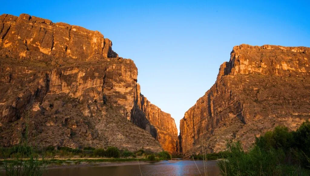 View of the steep walls of Santa Elena Canyon with a river running through the canyon and vegetation on the right at sunset when the canyon looks golden. 