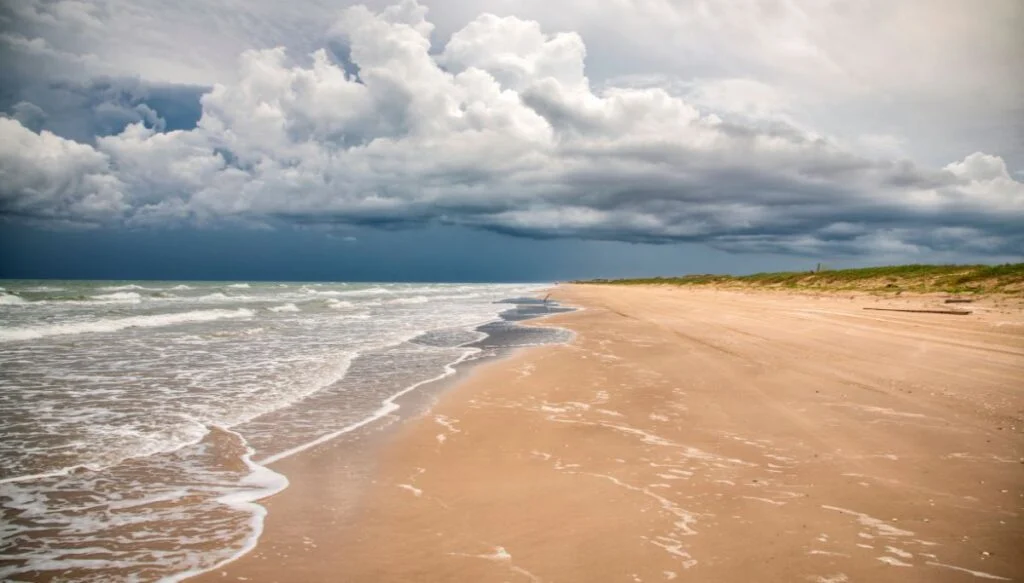 View of water crashing up against the sandy shore of a beach on South Padre Island. The sky is cloudy and there is grass along the edge of the sand. 