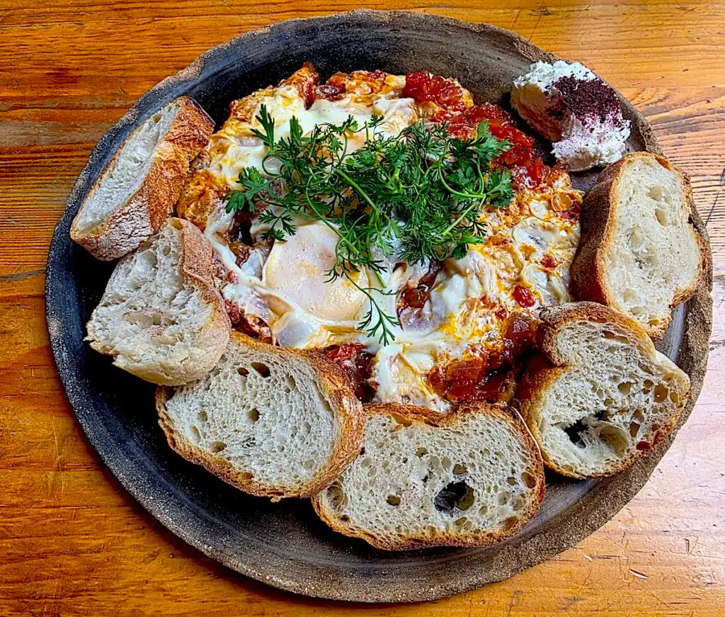 Aerial view of Shakshuka surrounded by bread with cheese on the side from Boulenc. 