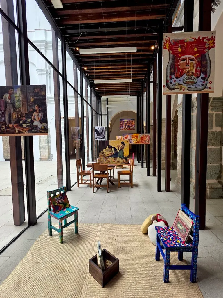 View of a display in Centro Cultural San Pedro with tables and chairs and paintings hanging in the air. 