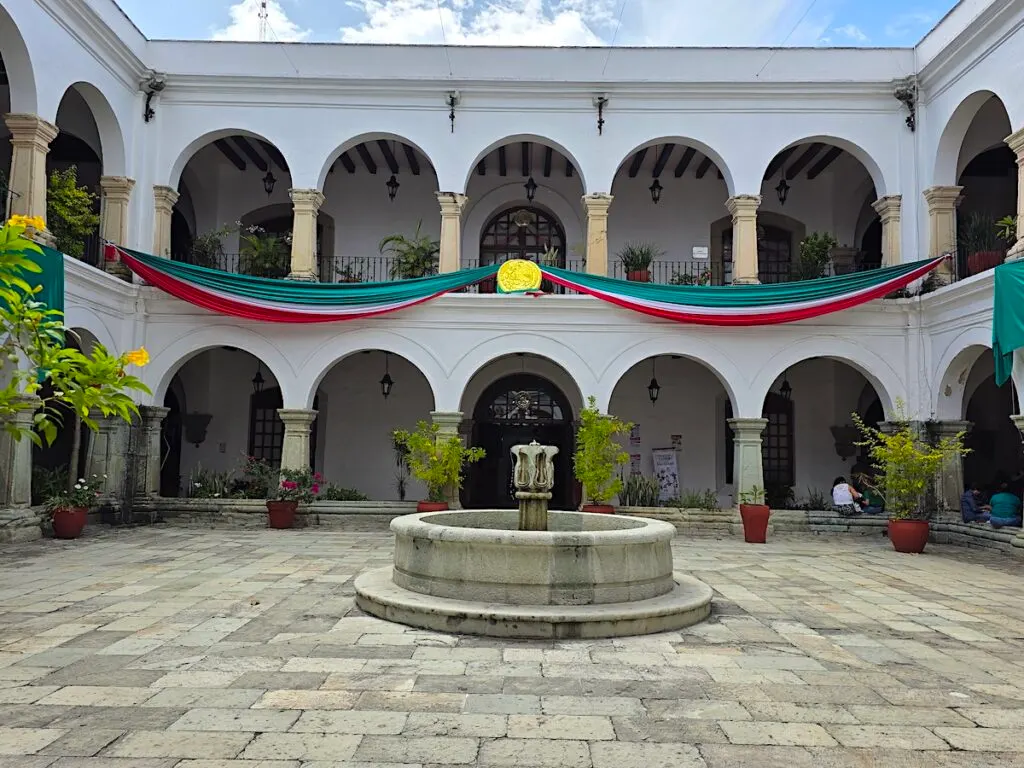View of a convent with arches and a central fountain. You can also see green, white, and red streamers hanging from the second floor. 