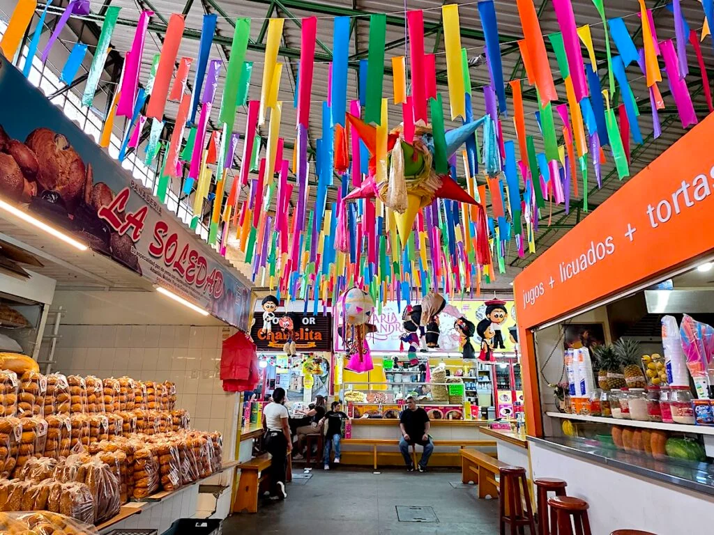 View of a pinata and colorful streamers hanging from the ceiling in Mercado 20 de Noviembre. You can see stalls selling food and people sitting and eating. 