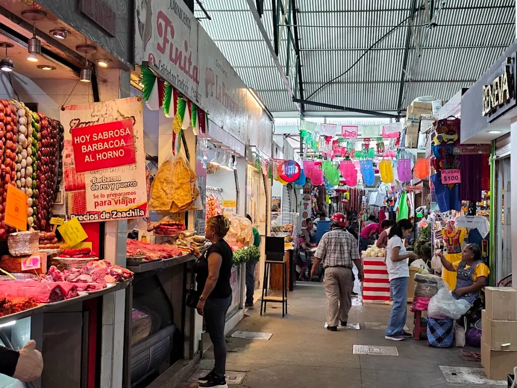 View of people walking through Mercado Benito Ju&aacute;rez with vendors selling meat on one side and people chilling on the other. You can also see colorful flags hanging in the air. 