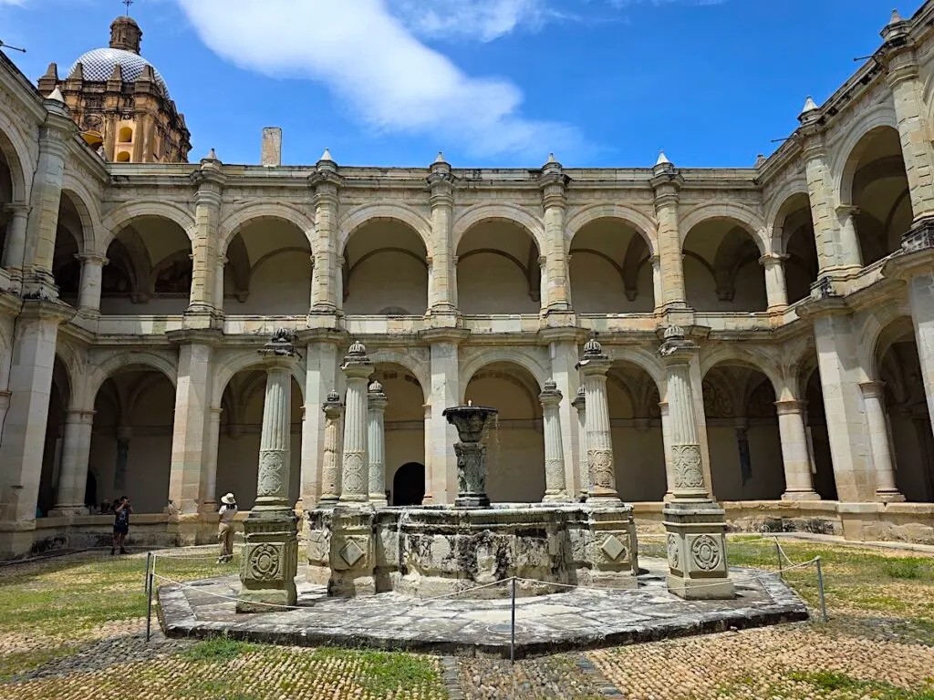 View of a central fountain inside a courtyard surrounded by stone archways with the steeple of a church on the left on a sunny day. 