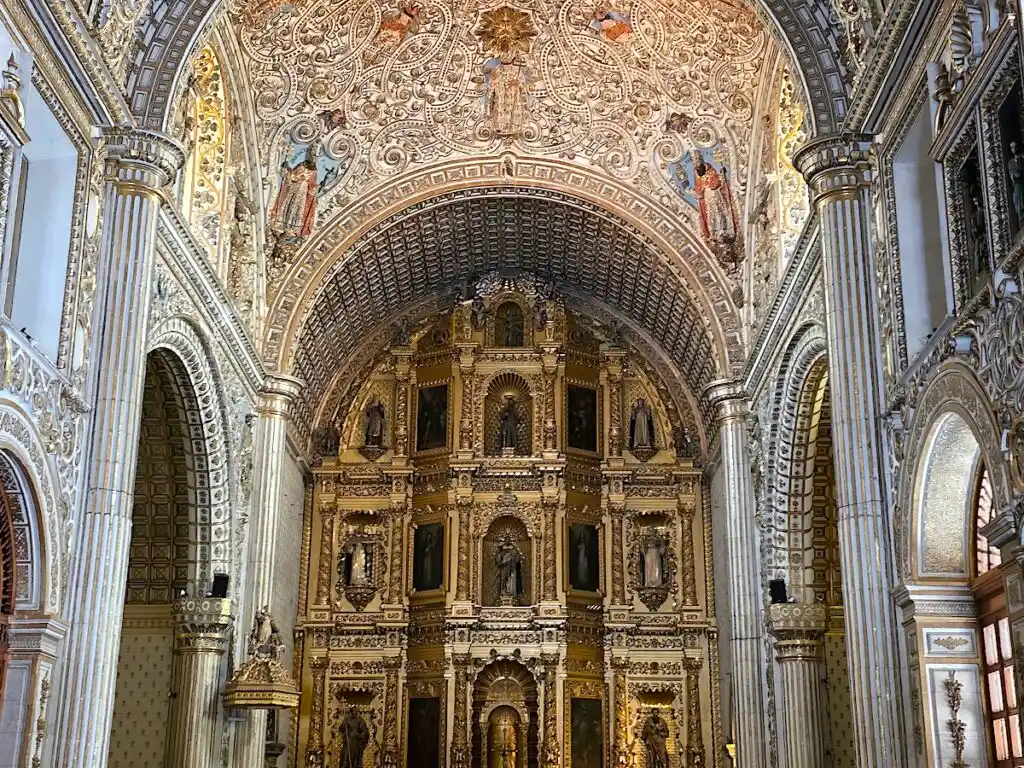 View of the interior of the Templo de Santo Domingo with its ornate ceiling and altar area with gold and lavish portraits of saints. 