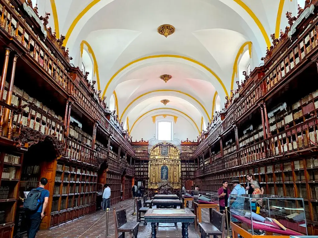 View of the interior of Biblioteca Palafoxiana. You can see an ornate golden wall with a saint at the end of the room and wooden shelves filled with books lining the walls. This is one of the best things to do in Puebla, Mexico. 