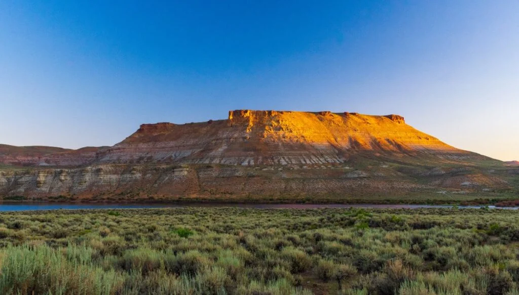 View of flat topped mountains that are turning gold in the light with a river at the base and vast meadows in the foregrounf. 