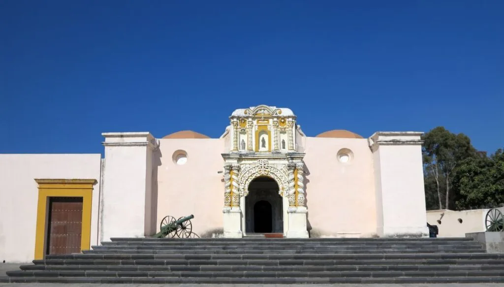 View of the white, Colonial-style exterior of Uerte de Loreto in Puebla. You can see a small cannon to the left and on top of the stairs. 
