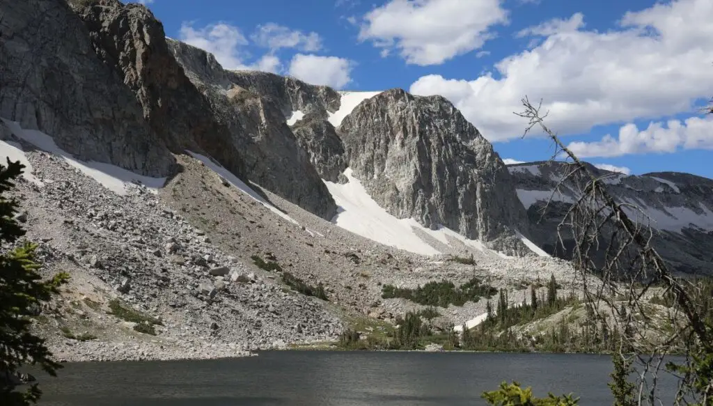 View of gray rock mountains with snow all over them and a blue sky in the background with a river in front of the mountains. 