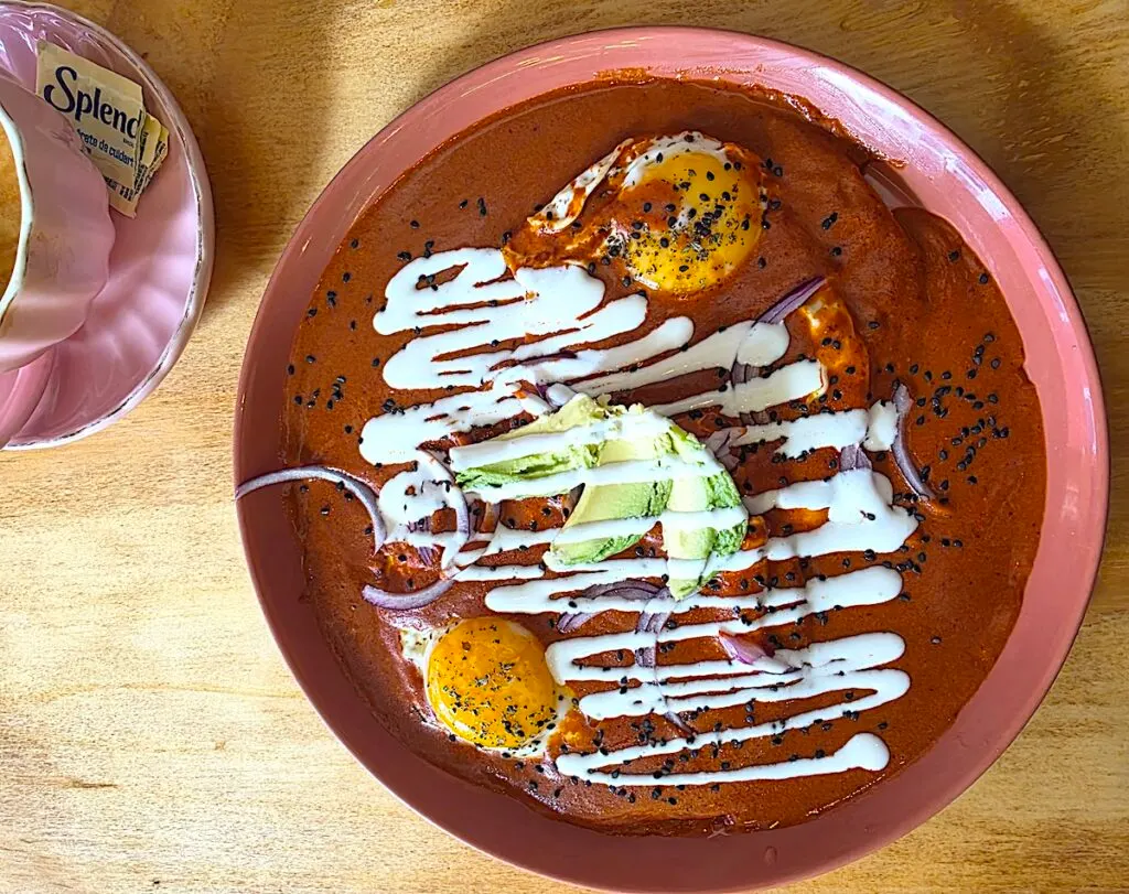 Aerial view of two eggs on tops of tortillas with dark orange pipian sauve on a pink plate with a pink cup on the left side. 