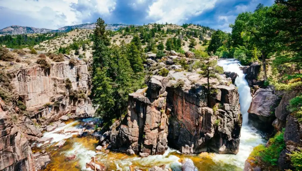 You see the waterfall on the right and a river surrounding an island of rock covered in Pine trees with pine tree covered mountains in the background. 