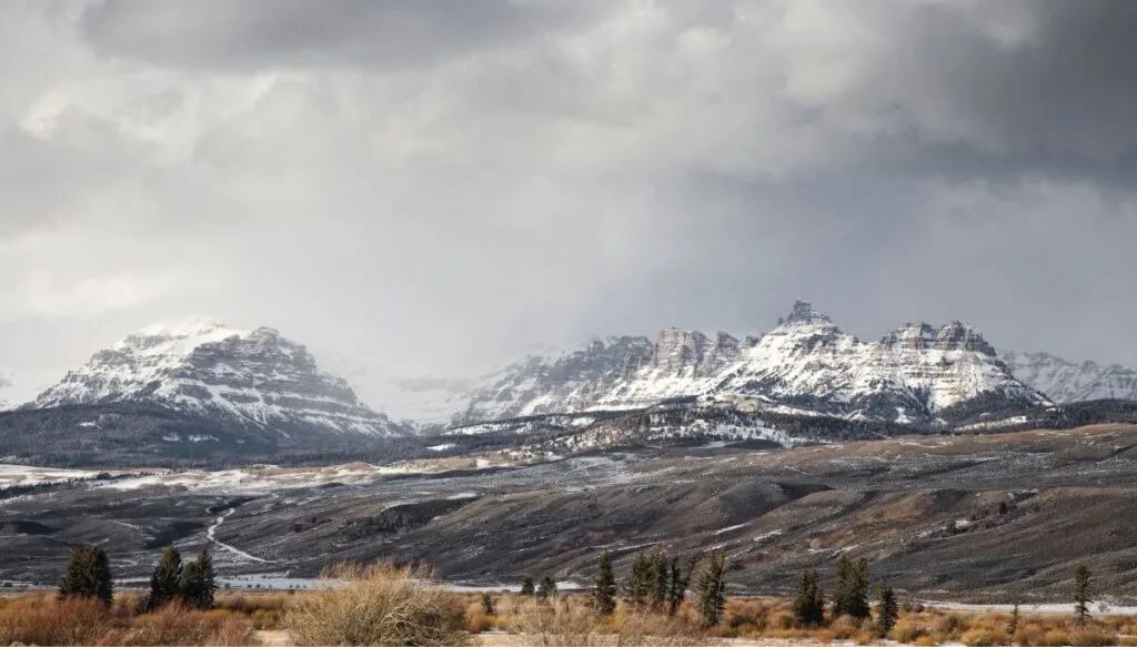 View of the snow covered mountains of the Togwotee Pass and a road going past the base of the mountains on a cloudy day. 