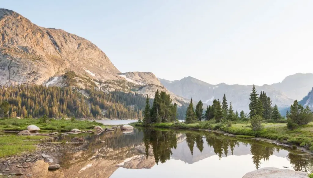 View of gray, rocky mountains with a lught dusting of snow and a base of pine trees with a  still body of water in the foreground. 