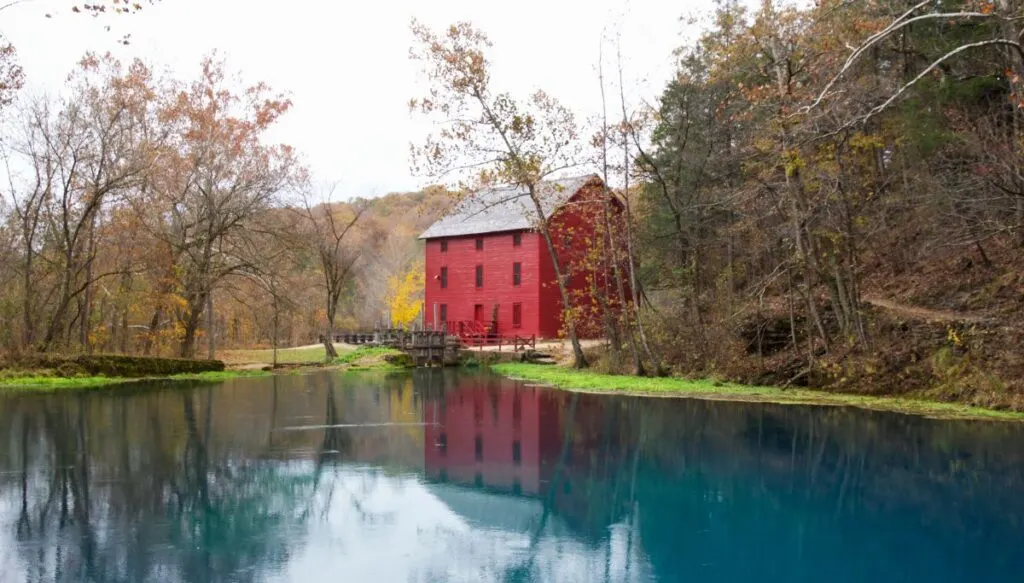 View of a vibrant red building sitting along a river filled with bright blue water that is surrounded by trees changing color and losing their leaves in the fall. This is Alley Spring and Mill and it is one of the most beautiful places in Missouri.