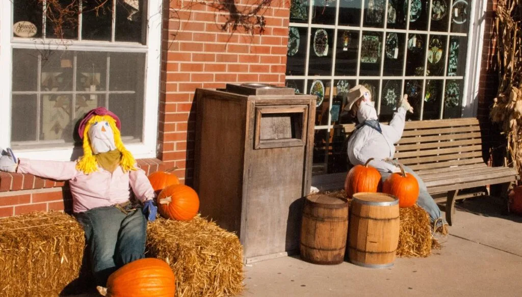 View of scarecrows sitting on hay bails and beside pumpkins on a street in Amana Colonies. 