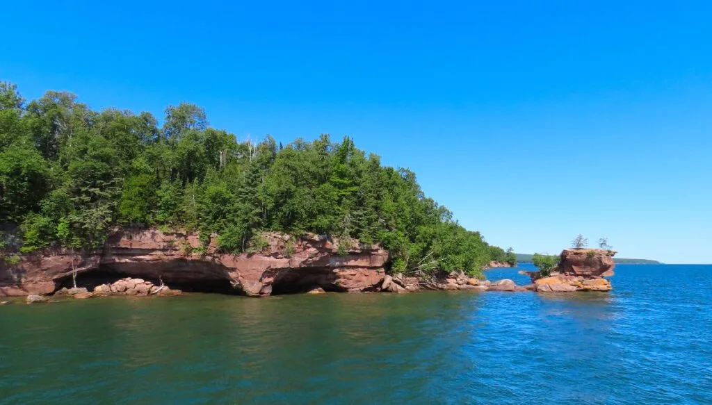 View of rugged red rocks sitting in the blue water with trees growing on top of the rocks. 