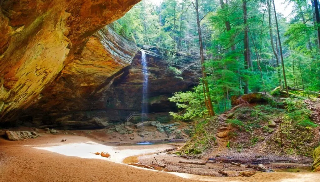 View from the inside of Ash Cave. You can see a small waterfall coming down from the roof of the cave on the left and a series of tall green trees on the right. 