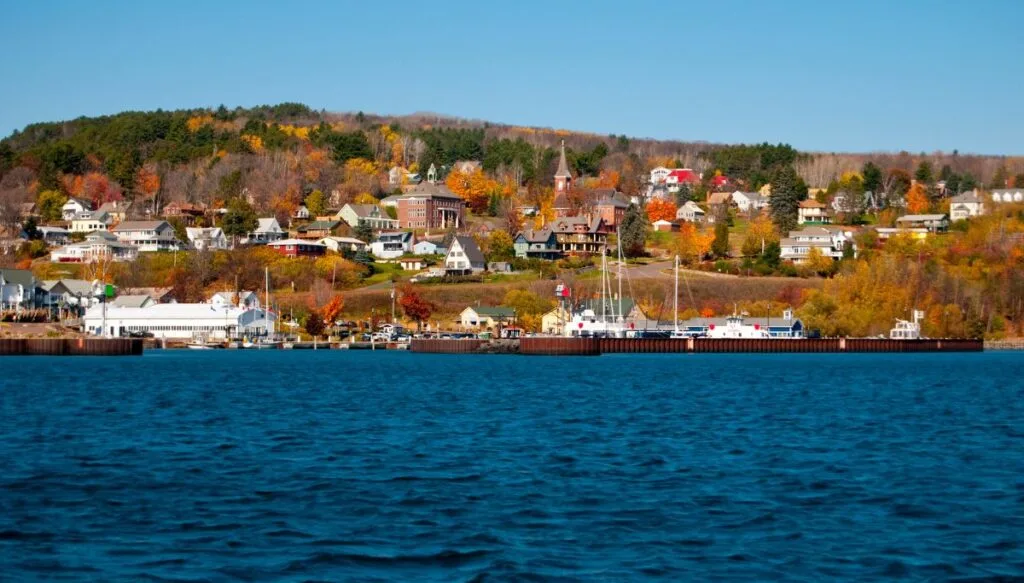 View of homes along the shore and ships docked along the shore of the island. There is also a ton of fall foliage surrounding the homes on the island. 