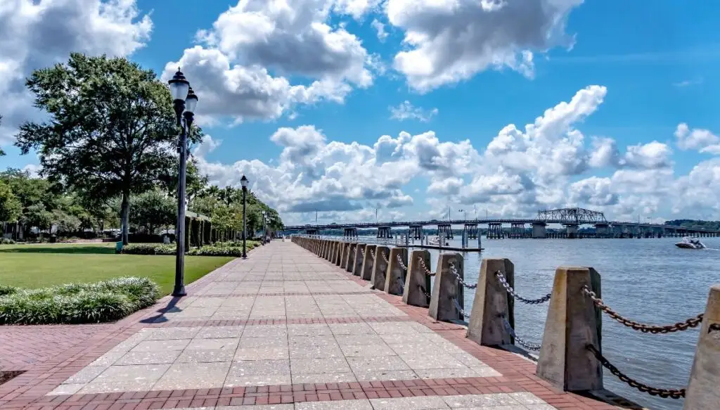 View of the waterfront in Beaufort. You can see a paved walkway lined with trees and a bridge in the background. The water is on the right and there are clouds in the sky. 