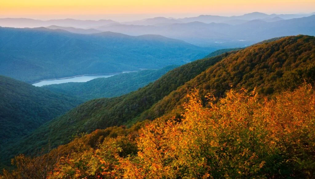 Panoramics view of colorful fall foliage and sweeping green forests from an overlook along the Blue Ridge Parkway with the sun setting in the background. 