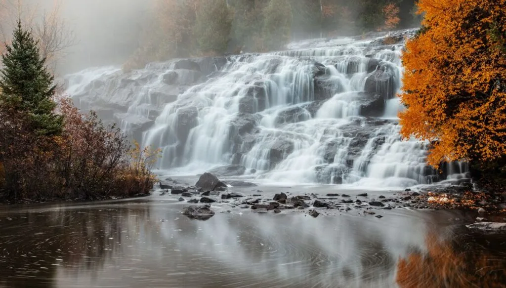 View of Bond Waterfall with a tree with vibrant orange foliage on the right. You can also see a quiet river at the base with some small rocks in it. 