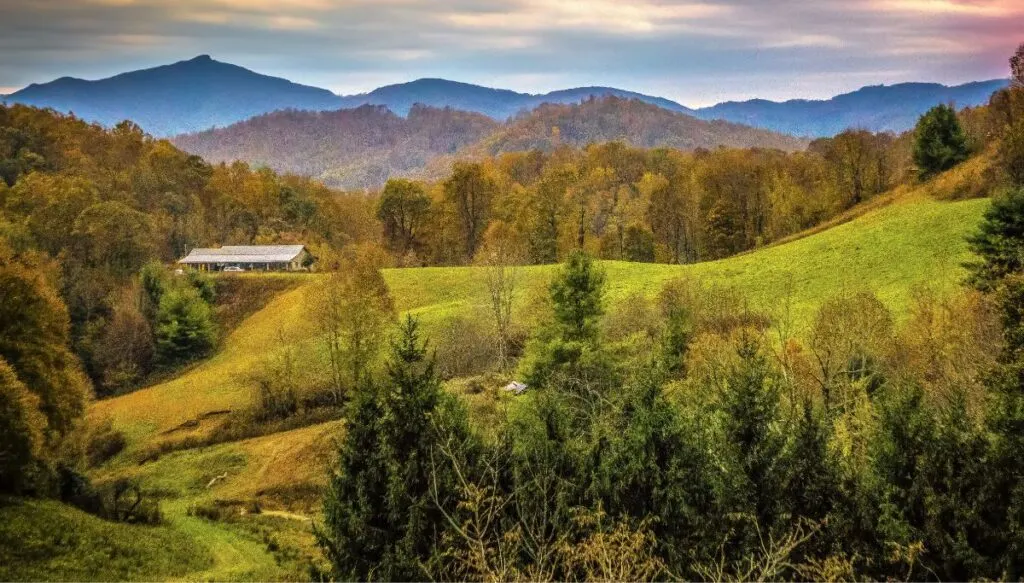 View of a farmhouse in a green meadow with mountains all around it in Boone, North Carolina. 