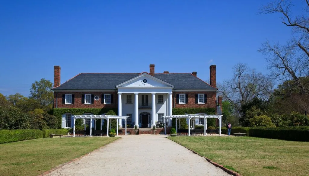 View of a brick home with white columns in the front and lawns on wither sida of a dirt road in the center. 