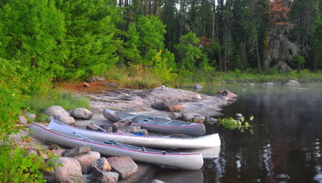View of canoes resting against the rocky shore of Boundary Waters Canoe Area Wilderness with green trees in the background.