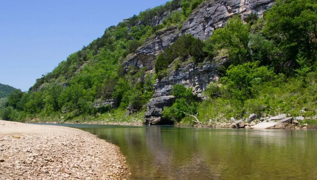 View of the Buffalo River in Arkansas. You can see a hill on the right full of green trees, the river in the middle, and a rocky bank on the left. 