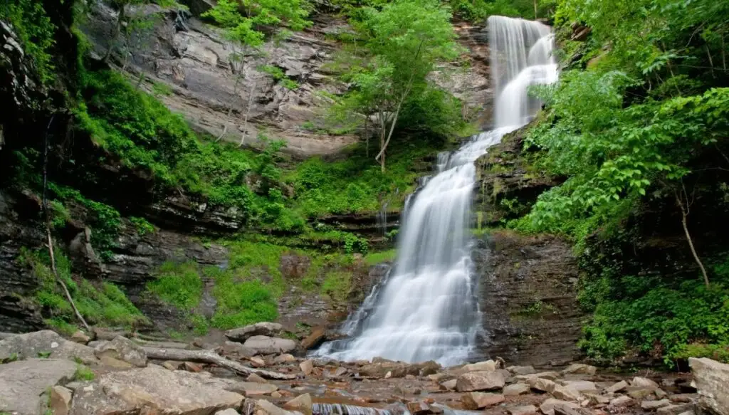 View of a waterfall falling down a hillside with greenery all around it and rocks sitting at the base of the waterfall. 