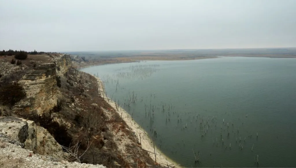 View of the blue water of Cedar Bluff Reservoir on the right and the rocky shores of the reservoir on the left. You can also see tall grass poking out of the water and sand lining the shores of the reservoir. 