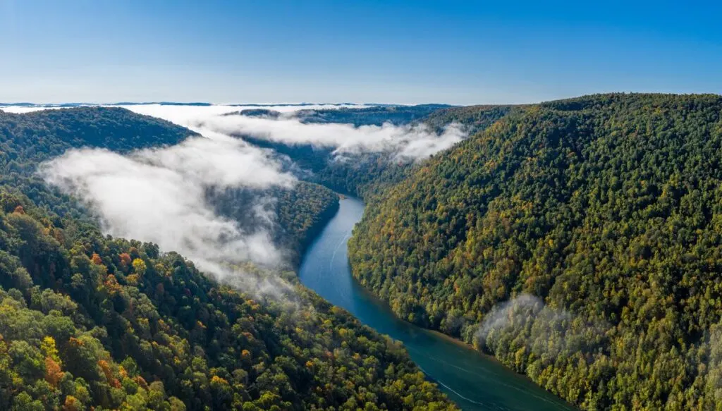 Aerial view of the blue Cheat River snaking past hills full of trees with mist hanging in the air on a sunny day. 