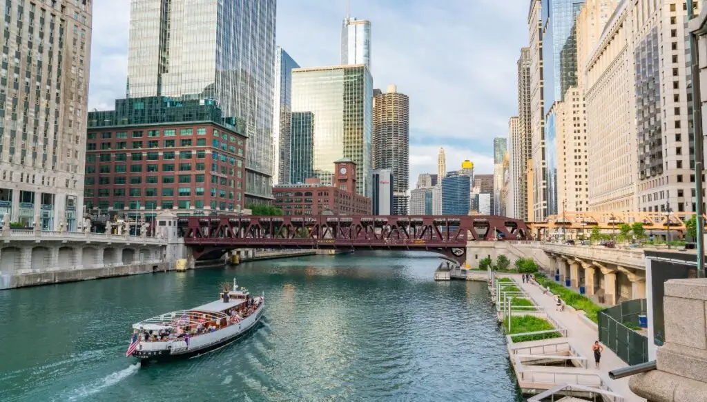 View of the river in Chicago with a boat going through the water and a sidewalk with people walking on the right. 