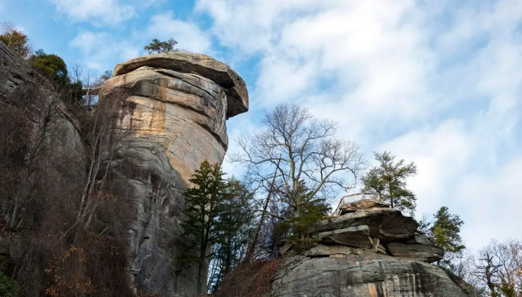 View of a circular shaped rock that looks like a chimney and that is surrounded by bare trees and pine trees with a blue sky in the background. 