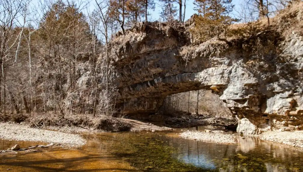 View of a natural stone bridge going over a stream in the winter since you can see trees without leaves all around the bridge. 