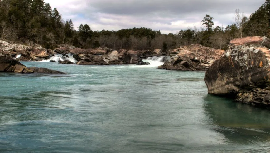 View of the blue Cossatot River with tons of rocks all along it. You can see tall trees in the background as well as two small waterfalls. 